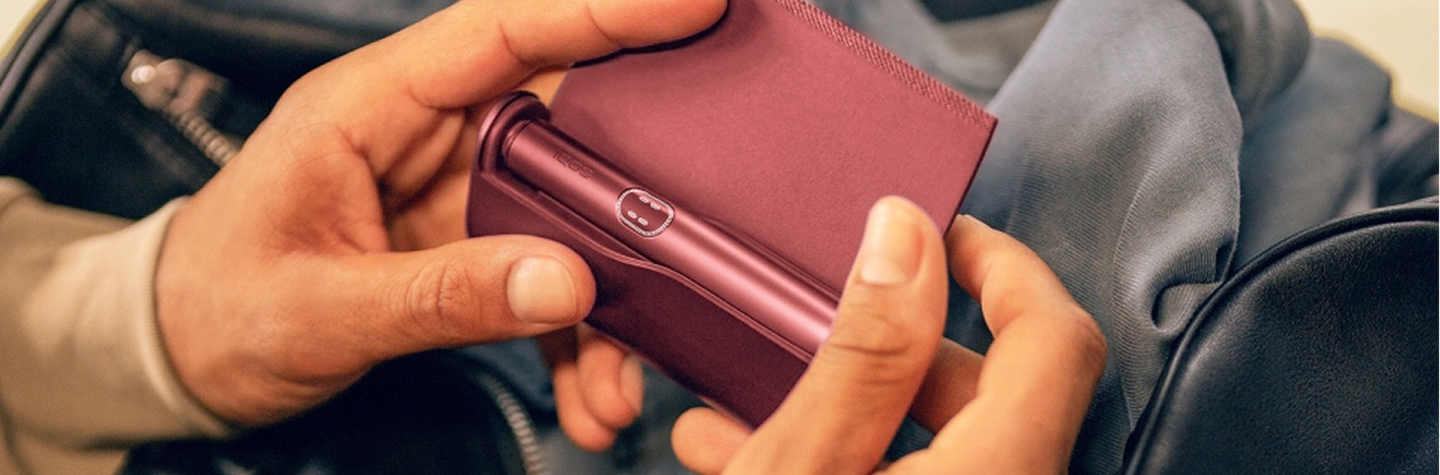 Руки держат IQOS ILUMA i PRIME над сумками.